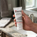 A hand holding a white squeeze tube of Stratus Performance Facial Moisturizer, part of a post workout skincare routine, in front of a blurred bathroom sink background.