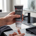 A hand holds a squeeze tube of Stratus Performance Facial Cleanser over a bathroom sink, to clean the face as part of a post workout skincare routine