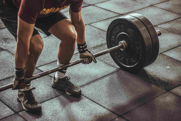 A close-up action shot of a person lifting a barbell with black weight plates on a dark gym floor.